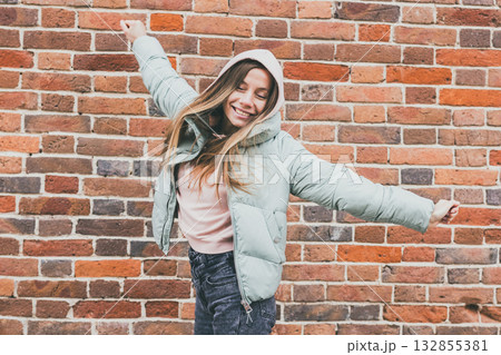 Joyful woman in pastel jacket raising arms and smiling outdoors. Urban freedom, positive energy, and expression of happiness in city life. 132855381