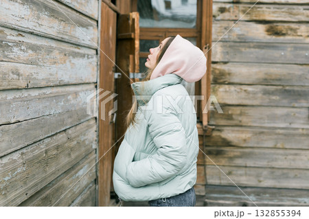 Young woman in mint jacket with pink hood looking up near vintage wooden house. Reflection, calm mood, and thoughtful atmosphere in urban winter setting. 132855394