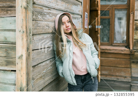 Woman in mint puffer jacket with hood standing near old wooden house. Winter atmosphere, calm confidence, and natural urban simplicity. Woman in mint puffer jacket with hood standing near old wooden house. Winter atmosphere, calm confidence, and natural urban simplicity. 132855395