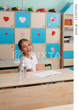 Smiling girl sitting at classroom desk with notebook and sanitizer at school. Joy, optimism, and positive atmosphere in modern education. 132855402