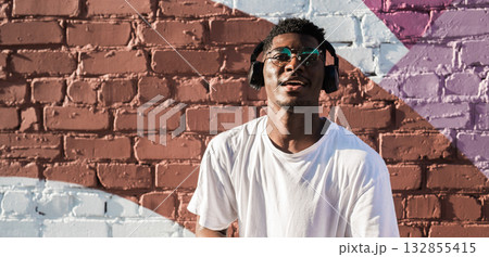 Young african american man dancing with headphones on colorful wall background. Concept of music, joy, freedom and urban lifestyle. Copy space banner 132855415