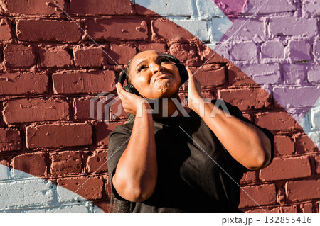 African american woman dancing playfully with headphones against painted wall. Concept of joy, energy, fun and youth lifestyle. African american woman dancing playfully with headphones against painted wall. Concept of joy, energy, fun and youth lifestyle. 132855416