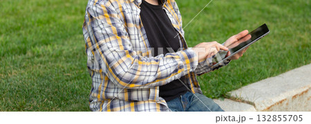 Man sitting on grass using tablet for work or study in sunny outdoor environment. Digital freedom, remote work, and mobile lifestyle. 132855705