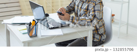 Male freelancer using smartphone while working on laptop at white desk. Banner with copy space. Multitasking, communication, and modern digital work concept. 132855706