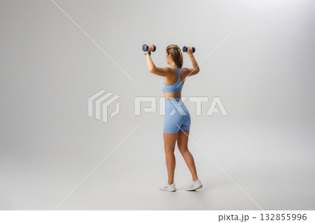 Fit woman exercises with dumbbells in stylish blue athletic wear indoors during bright lighting session Fit woman exercises with dumbbells in stylish blue athletic wear indoors during bright lighting session 132855996