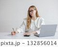 Young woman working at desk with laptop and calculator while taking notes in a bright, modern workspace during the day 132856014