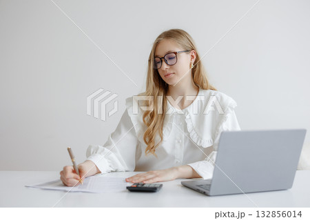 Young woman working at desk with laptop and calculator while taking notes in a bright, modern workspace during the day 132856014