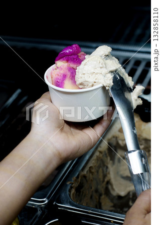 Close-up of a hand scooping colorful ice cream into a cup at a gelato shop, showing vibrant pink and beige flavors, stainless steel scoop, and creamy texture in a realistic preparation process scene 132858110