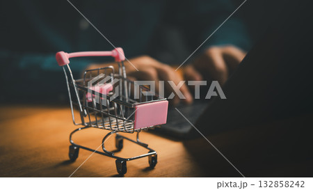 Shopping cart on desk with person using laptop. Concept of online shopping, digital marketplace, e-commerce website, product search, checkout process, retail marketing, consumer purchasing behavior. 132858242
