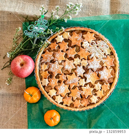 Homemade Christmas pie topped with star shaped cookies and powdered sugar, festive holiday dessert on rustic table with apple, oranges, and Merry Christmas decoration, cozy winter baking atmosphere 132859953