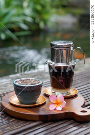 Vietnamese drip coffee with condensed milk and ceramic cup on wooden board, tropical frangipani flower, outdoor cafe scene, authentic slow coffee lifestyle and warm natural atmosphere 132860570