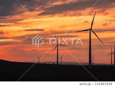 Multiple wind turbines silhouetted against a vibrant sunset sky. The turbines stand tall against the backdrop of orange and red clouds. 132861938
