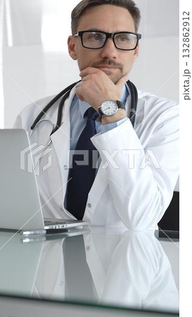 Male doctor wearing spectacles, thinking about healthcare diagnosis while using a laptop in a clinic office. Medicine and health care Male doctor wearing spectacles, thinking about healthcare diagnosis while using a laptop in a clinic office. Medicine and health care 132862912