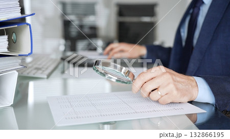 Professional male hand holding magnifying glass, looking at financial data on paper, checking details in accounting and finance. Topic of audit and taxes in business 132866195