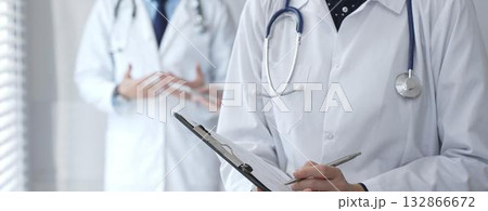 A medical professional with a stethoscope writes on a clipboard while a doctor and female assistant collaborate in a clinic, discussing patient data and healthcare information A medical professional with a stethoscope writes on a clipboard while a doctor and female assistant collaborate in a clinic, discussing patient data and healthcare information 132866672