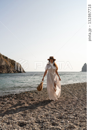 Woman, beach, vacation: elegant woman in white dress walks on pebble shore with straw bag, serene summer travel copy space 132867718