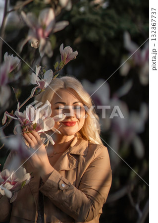A woman is holding a magnolia flower in her hand and standing in front of a tree. Concept of serenity and beauty, as the woman is surrounded by nature and the flower adds a touch of color. 132867737