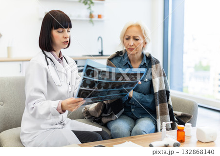 A female doctor in a white coat reviews an X-ray with an elderly woman wrapped in a blanket, discussing medical results. 132868140
