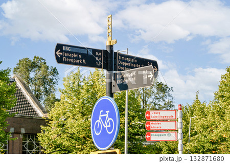 Multiple directional signs indicate various routes and locations, featuring cycling symbols and distances to nearby areas, under a clear sky. 132871680