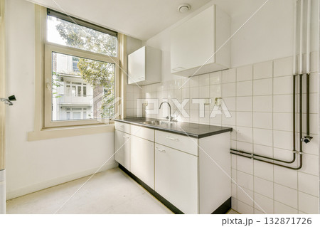 A clean, contemporary kitchen featuring white cabinets, a stainless steel countertop, and large windows bringing in natural light. 132871726