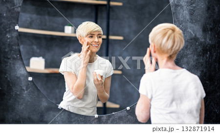 Woman Applying Facial Cream Moisturizing Aging Skin Smiling To Her Reflection In Mirror Standing In Bathroom Indoor. Selective Focus Woman Applying Facial Cream Moisturizing Aging Skin Smiling To Her Reflection In Mirror Standing In Bathroom Indoor. Selective Focus 132871914