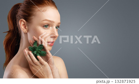 Profile Headshot Of Ginger-Haired Young Lady Holding Exotic Green Plant Near Face Posing Standing Over Gray Studio Background, Looking Aside At Copy Space. Natural Cosmetics Advertisement 132871925