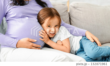 Cute little daughter listening to expecting mother belly, sitting on couch, close up Cute little daughter listening to expecting mother belly, sitting on couch, close up 132871961