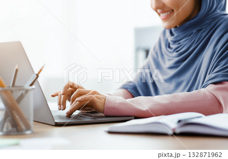 Unrecognizable muslim woman in hijab working on laptop computer in office, typing on keyboard and smiling, cropped image with empty space 132871962