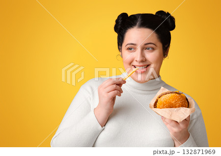 Brunette plump girl eating tasty hamburger and french fries, enjoying fast food and looking aside at copy space over yellow background, panorama Brunette plump girl eating tasty hamburger and french fries, enjoying fast food and looking aside at copy space over yellow background, panorama 132871969