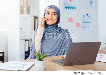 Business Communication. Joyful Arabic Female Employee Talking On Cellphone And Using Laptop While Working In Modern Office 132871975
