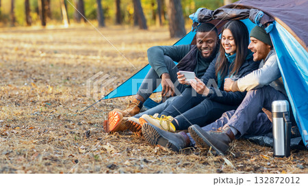 Smiling multiethnic backpackers looking at smartphone, drinking tea, camping together at autumn forest, empty space Smiling multiethnic backpackers looking at smartphone, drinking tea, camping together at autumn forest, empty space 132872012