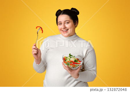 Dieting Concept. Overweight girl holding bowl with fresh vegetable salad, eating healthy food, posing over yellow background in studio, panorama Dieting Concept. Overweight girl holding bowl with fresh vegetable salad, eating healthy food, posing over yellow background in studio, panorama 132872048
