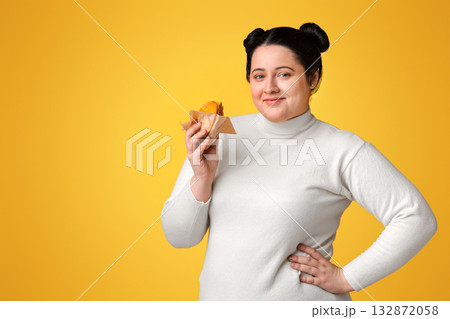 Beautiful plump woman posing with burger in hand over yellow background and smiling at camera, empty space 132872058