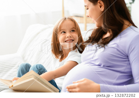 Close up of little girl looking at pregnant mom while reading book at home, free space Close up of little girl looking at pregnant mom while reading book at home, free space 132872202