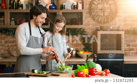 Young father and daughter cooking meal together, adding seasoning to salad, enjoying spending time in kitchen 132872203