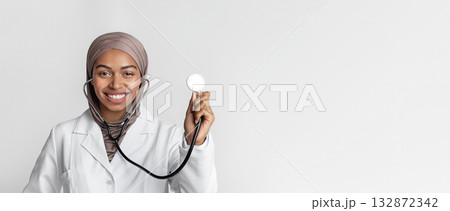 Portraif of young black muslim female doctor in hijab with stethoscope, posing over light background and smiling at camera, free space Portraif of young black muslim female doctor in hijab with stethoscope, posing over light background and smiling at camera, free space 132872342