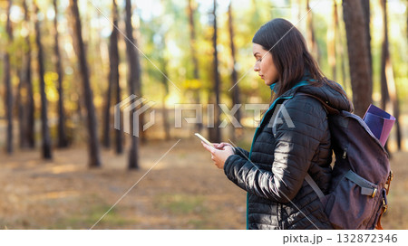 Side view of serious girl with backpack using mobile phone in forest, checking location, copy space Side view of serious girl with backpack using mobile phone in forest, checking location, copy space 132872346