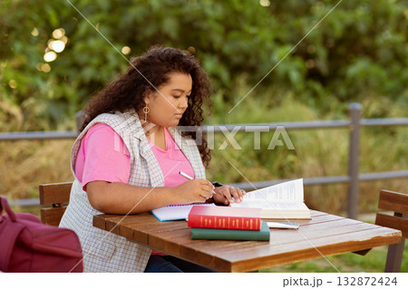 A student engaged in studying while seated at a wooden table outdoors. The individual writes in a notebook, with books beside them, immersed in their studies on a sunny day. 132872424