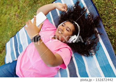 A young lady relaxes on a striped blanket in a grassy park, wearing headphones and holding a smartphone. She smiles while enjoying her music under the warm sun. A young lady relaxes on a striped blanket in a grassy park, wearing headphones and holding a smartphone. She smiles while enjoying her music under the warm sun. 132872442