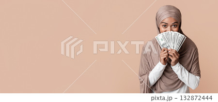 Happy Black Muslim Woman In Headscarf Covering Face With Dollar Cash Fan, Standing Over Yellow Background, Panorama With Copy Space 132872444