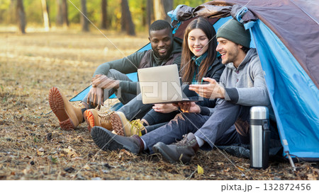 Happy international friends sitting in tent, using laptop and talking, forest background, copy space Happy international friends sitting in tent, using laptop and talking, forest background, copy space 132872456