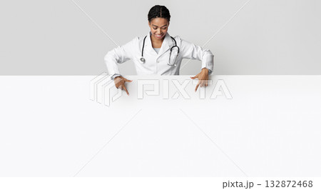 Medical offer. Afro female doctor looking down and pointing at blank white advertising board with empty space for text 132872468
