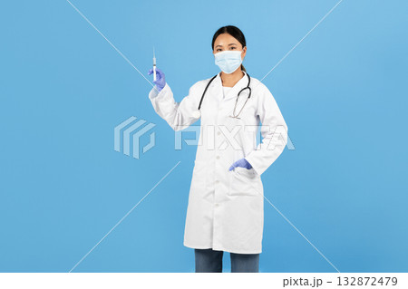 A healthcare worker, dressed in a white lab coat and blue gloves, confidently holds up a syringe while wearing a surgical mask, set against a vibrant blue backdrop. 132872479