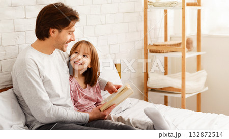Loving dad reading fairy tales to his little curious daughter, sitting on bed together 132872514