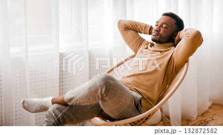 Weekend. Relaxed African American Man Sitting On Modern Chair Holding Hands Behind Head Next To Window At Home. Selective Focus Weekend. Relaxed African American Man Sitting On Modern Chair Holding Hands Behind Head Next To Window At Home. Selective Focus 132872582