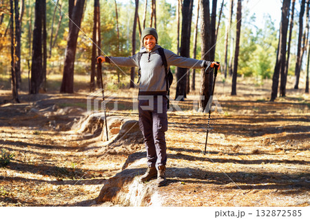 Cheerful guy backpacker with walking sticks posing in sunlight at autumn forest 132872585