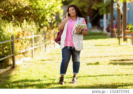 A young woman stands in a green park, talking on her phone while carrying books. She wears a bright pink shirt and a long vest, enjoying the sunny day surrounded by greenery. A young woman stands in a green park, talking on her phone while carrying books. She wears a bright pink shirt and a long vest, enjoying the sunny day surrounded by greenery. 132872591