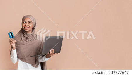 Online payments concept. Thoughtful black islamic girl in headscarf with laptop and credit card and looking away, yellow background with empty space Online payments concept. Thoughtful black islamic girl in headscarf with laptop and credit card and looking away, yellow background with empty space 132872647