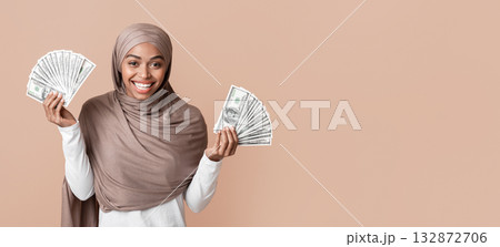 Lucky Winner. Overjoyed Black Muslim Girl in Hijab Holding Lots Of Dollar Cash In Both Hands, Standing Over Yellow Background With Free Space 132872706
