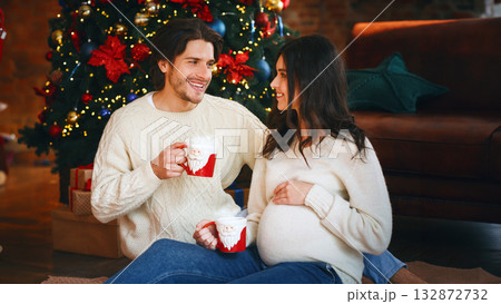 Young married couple drinking cocoa at home next to Christmas tree, having conversation, pregnant woman holding her belly, celebrating xmas together 132872732